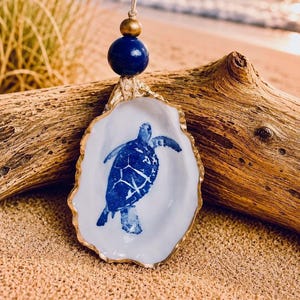 Puede incluir: Un adorno decorativo de concha de ostra con una ilustración de una tortuga marina azul en el interior. Los bordes de la concha están pintados de dorado y cuelga de un cordel de yute adornado con una cuenta azul marino grande y una cuenta dorada más pequeña.