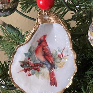 May include: A Christmas tree ornament depicting a painted cardinal bird on a white oyster shell with a gold-painted edge. The ornament is topped with a red bead and a gold bead, hanging on a green Christmas tree.