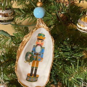 May include: A Christmas tree ornament featuring a painted oyster shell with a gold-painted rim. The shell displays a colorful nutcracker figure. A light blue bead and a gold bead are above the shell, suspended by a string.