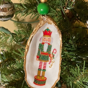 May include: A Christmas ornament made from a white oyster shell with a gold-painted edge. The ornament features a painted image of a nutcracker soldier in a green and red uniform, holding a candy cane. A green bead and gold bead are at the top.