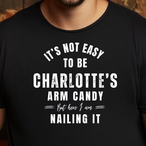May include: Black t-shirt with white text that reads "IT'S NOT EASY TO BE CHARLOTTE'S ARM CANDY - But here I am - NAILING IT." The shirt is worn by a person smiling at the camera.