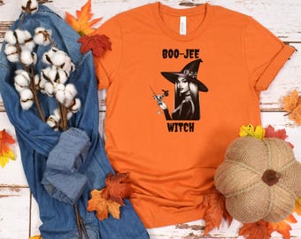 May include: Orange t-shirt with a black and white illustration of a witch holding a cocktail. The text "Boo-Jee Witch" is printed on the shirt.