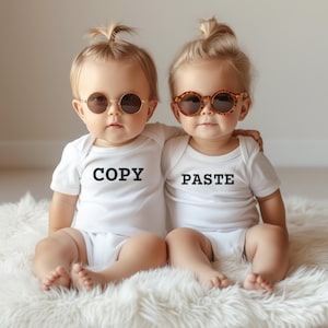 May include: Two babies wearing white onesies with the words "COPY" and "PASTE" printed on them. They are both wearing brown sunglasses and are sitting on a white fluffy surface.