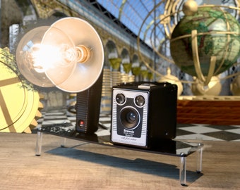 May include: A vintage black and silver Brownie camera with a flash attachment sits on a clear acrylic stand next to a table lamp with a clear glass shade and a light bulb. The camera is on a wooden surface with a black and white chequered pattern in the background.