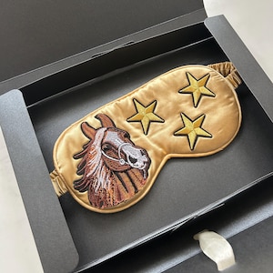 May include: A gold satin sleep mask featuring an embroidered horse head and three gold stars. The mask is displayed in an open black box, highlighting the design and texture of the sleep accessory.