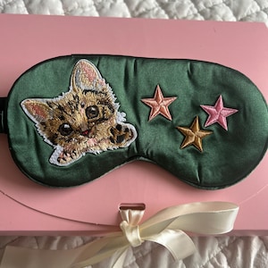 Peut inclure: Un masque de sommeil en satin vert avec un chat brodé et trois étoiles. Le chat est marron et beige avec des détails blancs. Deux étoiles roses et une étoile dorée sont brodées sur le masque. Le masque est sur une boîte rose avec un ruban crème.