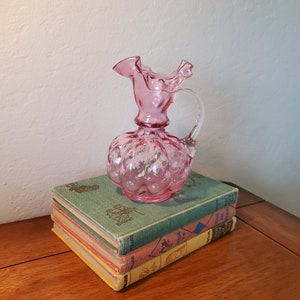 May include: A pink glass pitcher with a ruffled top and a clear handle. The pitcher has a textured, bubble-like design and is sitting on a stack of books.