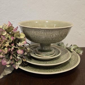 May include: A set of green ceramic tableware, including a large pedestal bowl and several plates, arranged on a dark wooden surface. The bowl and plates feature a detailed embossed pattern. Dried flowers are placed to the left of the tableware.