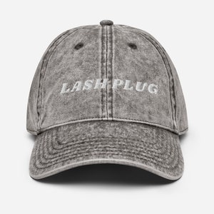 Pode incluir: Boné de beisebol cinza desgastado com o texto "LASH PLUG" bordado em branco. O boné tem uma aba curva e um botão na parte superior. Estilo vintage.