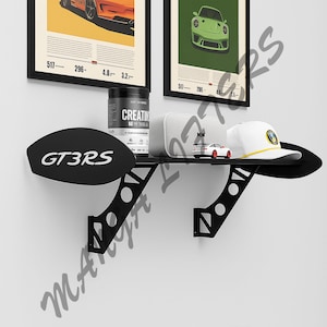 May include: A black metal shelf in the shape of a car spoiler, mounted on a white wall. The shelf holds a white hat, a small white and red toy car, a white speaker, and a black container. Two framed car posters are above the shelf.