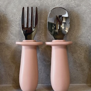 May include: A set of two pink handled baby utensils, a fork and a spoon, with stainless steel heads.