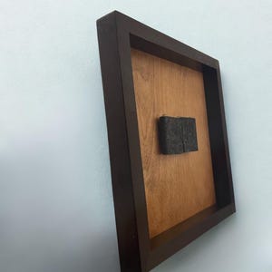 May include: A dark brown framed artwork with a light brown wooden interior and a dark gray rectangular object in the center. The frame is square and mounted on a light blue wall.