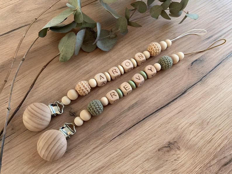 Dummy clip for babies, a handy accessory for parents to secure pacifiers to clothes, preventing loss. Crafted from natural wooden beads in various textures and neutral tones, some beads feature engraved letters forming names.