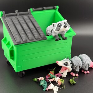 May include: A green toy bin with a black lid. Two small toy animals, a raccoon and a possum, are near the bin. The possum is eating colourful yarn.