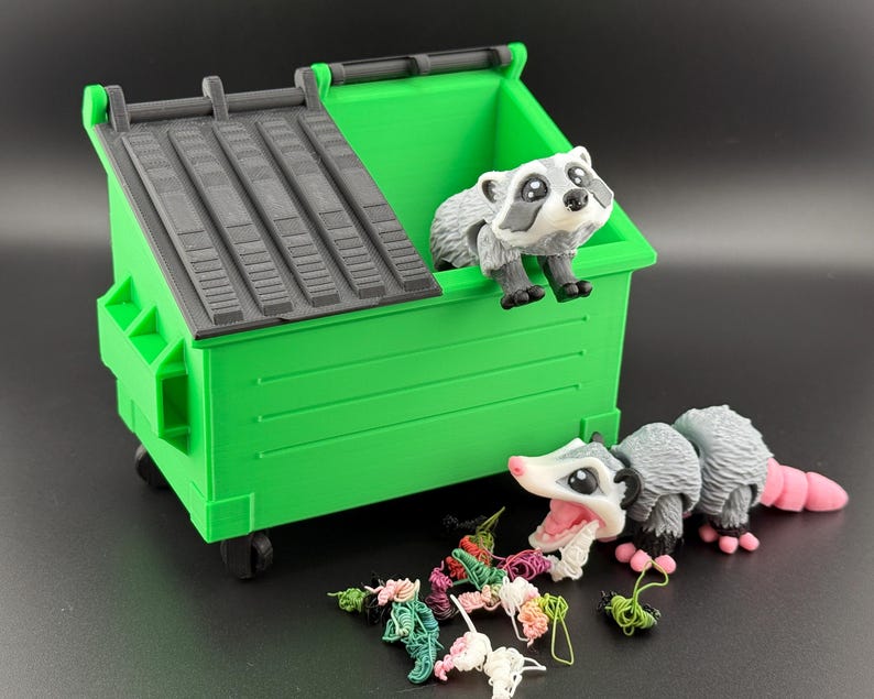 May include: A green toy bin with a black lid. Two small toy animals, a raccoon and a possum, are near the bin. The possum is eating colourful yarn.