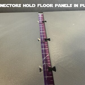 Puede incluir: Primer plano de dos paneles de suelo grises con una regla morada en el medio. Se ven conectores negros que sujetan los paneles. El texto "CONNECTORS HOLD FLOOR PANELS IN PLACE" est&aacute; en la parte superior.