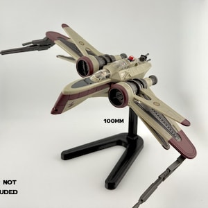 May include: A model starfighter in beige and burgundy, mounted on a black stand. The model has detailed wings and a cockpit, with a measurement of 100mm visible. The text "SHIP NOT INCLUDED" is also visible.