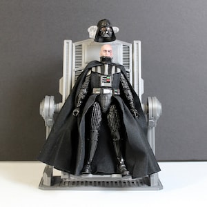 以下が含まれることがあります： グレーの玉座に座る黒いダース・ベイダーのアクションフィギュア。ヘルメットは取り外され、上に置かれています。フィギュアは黒いマントと鎧を着用し、胸部には詳細なパネルがあります。玉座は機械的なデザインです。