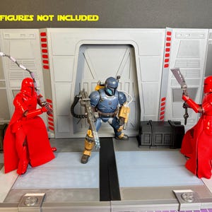 Puede incluir: Un diorama tem&aacute;tico de Star Wars con un interior gris y blanco y una puerta central. Dos figuras rojas con armas flanquean una figura blindada azul y amarilla. El texto "FIGURES NOT INCLUDED" se muestra en la parte superior.