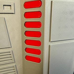 Puede incluir: Primer plano de un panel gris con una serie de siete botones rojos rectangulares. El panel tiene una superficie texturizada con varias formas geom&eacute;tricas y un recorte rectangular. El dise&ntilde;o parece ser un componente de una estructura m&aacute;s grande.