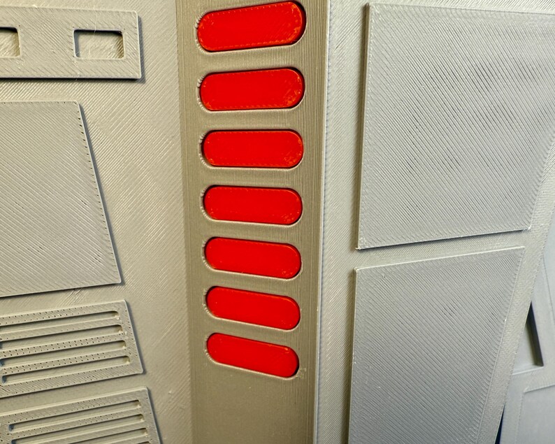 Puede incluir: Primer plano de un panel gris con una serie de siete botones rojos rectangulares. El panel tiene una superficie texturizada con varias formas geom&eacute;tricas y un recorte rectangular. El dise&ntilde;o parece ser un componente de una estructura m&aacute;s grande.