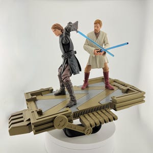 May include: Anakin Skywalker and Obi-Wan Kenobi action figures with blue lightsabers, standing on a detailed, tan and gray platform. The figures are posed in a battle stance, showcasing their outfits and accessories. The platform has a mechanical design.
