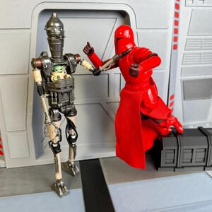 Puede incluir: Dos figuras de acci&oacute;n de Star Wars. Una es una figura de Guardia Imperial roja, y la otra es un droide con un compartimento que contiene una figura de Grogu. Las figuras est&aacute;n posadas en una escena con un fondo gris.