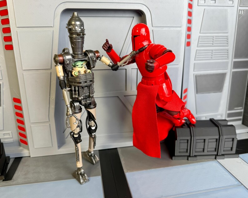 Puede incluir: Dos figuras de acci&oacute;n de Star Wars. Una es una figura de Guardia Imperial roja, y la otra es un droide con un compartimento que contiene una figura de Grogu. Las figuras est&aacute;n posadas en una escena con un fondo gris.