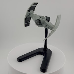 May include: A gray model of a TIE fighter spacecraft, a fictional starship from the Star Wars franchise, mounted on a black stand. The spacecraft has two solar panels and a central cockpit.