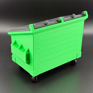 May include: A green plastic toy dumpster with black wheels and a black plastic lid. The dumpster has a textured surface and is designed to look like a real dumpster.