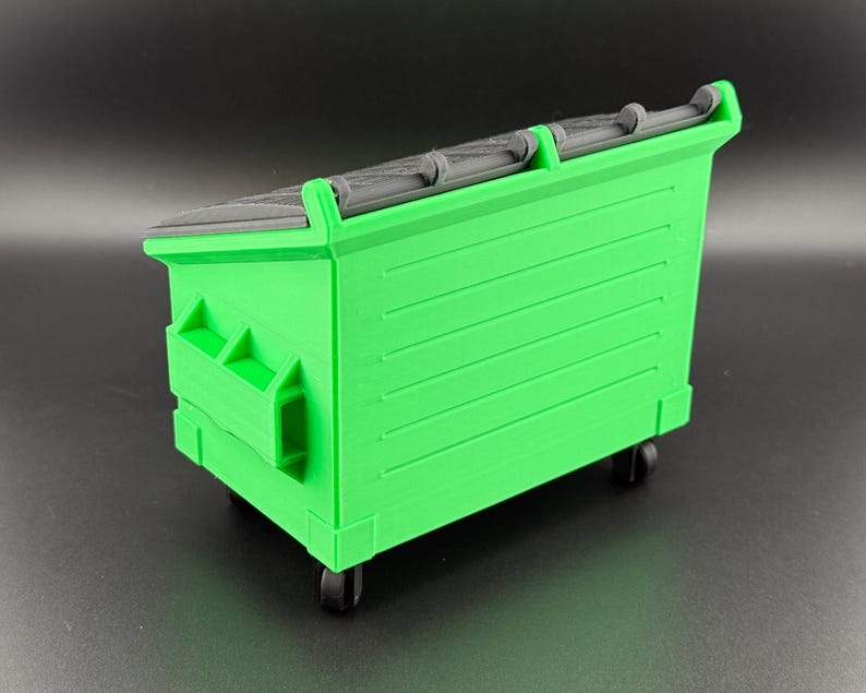 May include: A green plastic toy dumpster with black wheels and a black plastic lid. The dumpster has a textured surface and is designed to look like a real dumpster.