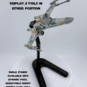 May include: A model X-wing starfighter in grey, green, and yellow, mounted on a black stand. The text "DISPLAY S FOILS IN EITHER POSITION" is above the starfighter. The text "ANGLE STANDS AVAILABLE WITH DYNAMIC PACK. ADJUSTABLE HEIGHT STANDS AVAILABLE WITH MULTI-HEIGHT PACK" is below.