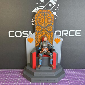 May include: A detailed action figure of a character seated on a throne. The throne is gray, orange, and red, with intricate geometric designs. The figure is wearing armor with blue and brown accents. The background features the text "COSMOS FORCE".