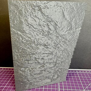 Puede incluir: Una pieza rectangular gris, texturizada, con un borde en capas. La superficie tiene una apariencia ondulada, similar al agua. El art&iacute;culo est&aacute; colocado sobre una esterilla de corte morada con l&iacute;neas de cuadr&iacute;cula blancas y un fondo negro.