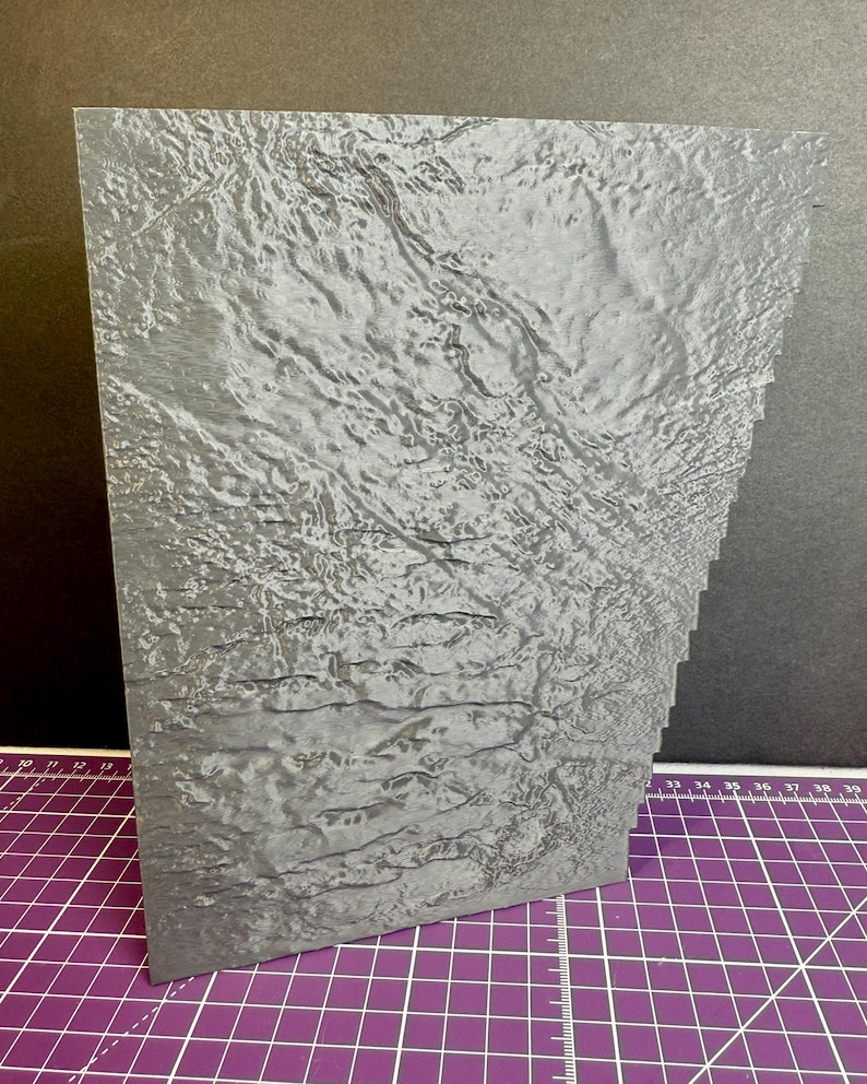 Puede incluir: Una pieza rectangular gris, texturizada, con un borde en capas. La superficie tiene una apariencia ondulada, similar al agua. El art&iacute;culo est&aacute; colocado sobre una esterilla de corte morada con l&iacute;neas de cuadr&iacute;cula blancas y un fondo negro.
