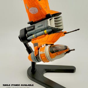 May include: A detailed model spaceship in orange and grey, mounted on a black stand. The spaceship features intricate wing designs and a cockpit. The stand has text: ANGLE STANDS AVAILABLE WITH DYNAMIC PACK. ADJUSTABLE HEIGHT STANDS AVAILABLE WITH MULTI-HEIGHT PACK.