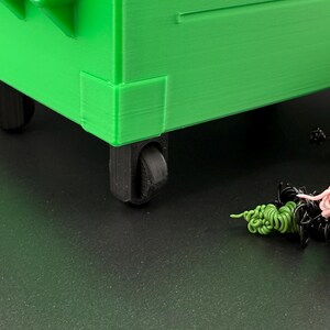 May include: A green plastic toy dumpster with black wheels. The dumpster is on a black surface with colourful strings of yarn.