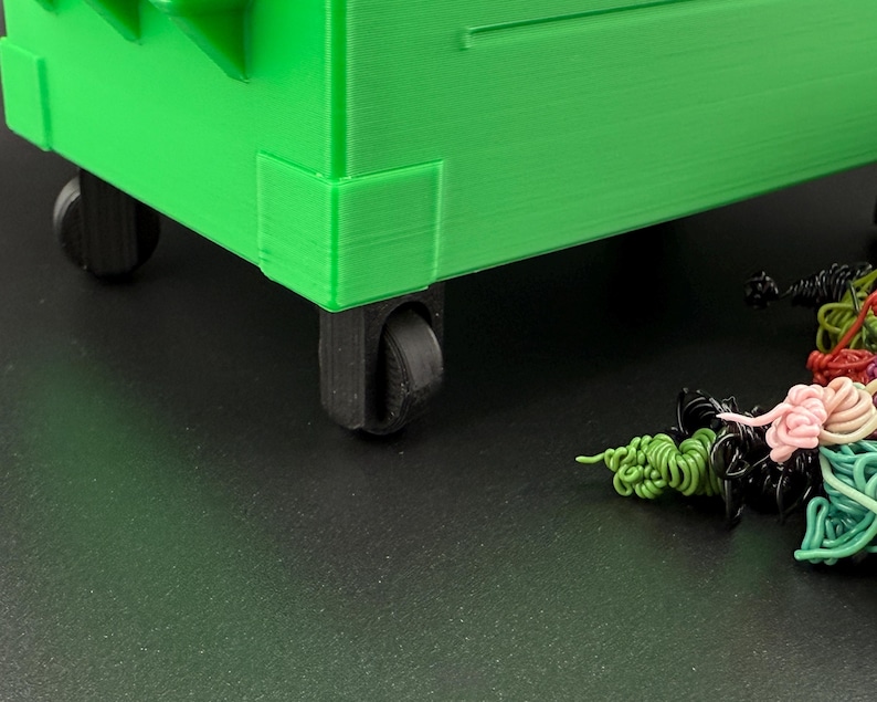 May include: A green plastic toy dumpster with black wheels. The dumpster is on a black surface with colourful strings of yarn.