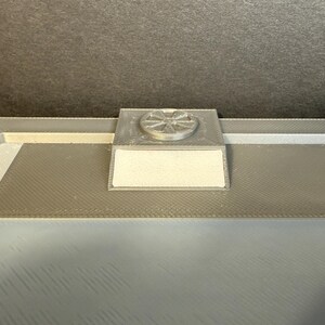 Puede incluir: Primer plano de una superficie rectangular gris y texturizada con un peque&ntilde;o objeto cuadrado plateado adjunto. El objeto plateado tiene un dise&ntilde;o circular en la parte superior. La superficie es de color gris claro y tiene un patr&oacute;n texturizado.