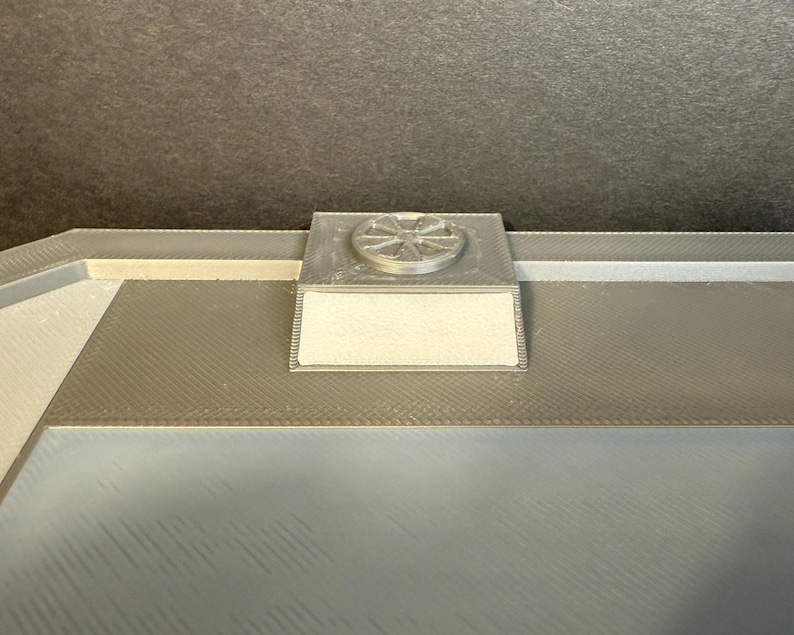 Puede incluir: Primer plano de una superficie rectangular gris y texturizada con un peque&ntilde;o objeto cuadrado plateado adjunto. El objeto plateado tiene un dise&ntilde;o circular en la parte superior. La superficie es de color gris claro y tiene un patr&oacute;n texturizado.