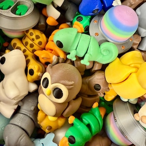 May include: A collection of colorful 3D printed figurines, including a chameleon, an owl, a penguin, a giraffe, and a spaceship. The figurines are made of plastic and have a variety of textures and finishes.