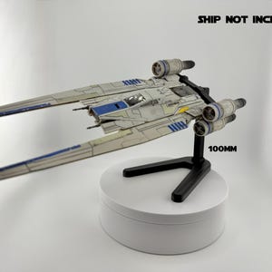 May include: A model starfighter in light gray, blue, and yellow, mounted on a black stand. The model is approximately 100mm in length. The text "SHIP NOT INCLUDED" is visible in the background.