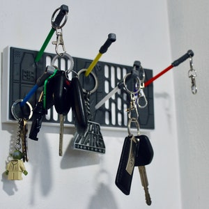 Könnte beinhalten: Ein weißer und grauer Schlüsselhalter im Star Wars-Design mit mehreren Schlüsseln und Schlüsselanhängern. Der Schlüsselhalter hat eine schwarz-weiße Grafik des Star Wars-Logos und den Text "Star Wars" darauf gedruckt. Zu den Schlüsselanhängern gehören eine Yoda-Figur, ein schwarzer und silberner Autoschlüssel und ein schwarzer und silberner Schlüssel mit einem silbernen Schlüsselanhänger.