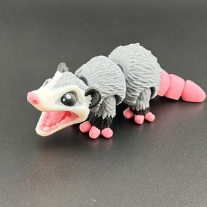 May include: A small, grey and white toy opossum with pink feet and a tail. The opossum has a wide open mouth and black eyes.