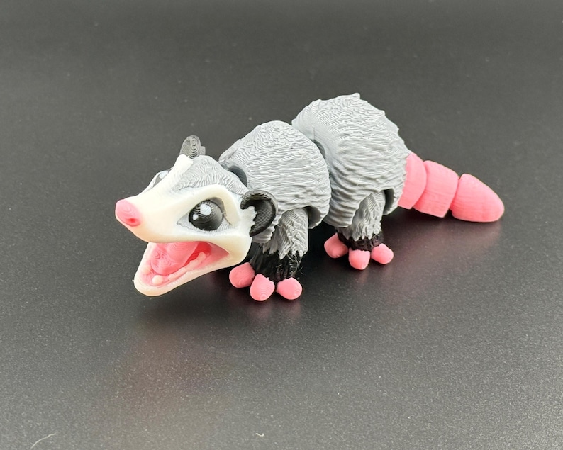 May include: A small, grey and white toy opossum with pink feet and a tail. The opossum has a wide open mouth and black eyes.