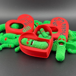 May include: Green pickle-shaped toys with red heart-shaped containers. The hearts have text that reads "You're a BIG dill" and "Pickle Love". The toys have smiling faces and are scattered on a dark surface.