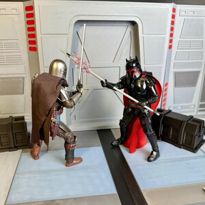 Puede incluir: Dos figuras de acci&oacute;n de Star Wars en una escena de batalla. Una figura es plateada y marr&oacute;n, con una capa y sosteniendo un bast&oacute;n blanco. La otra figura es negra y roja, tambi&eacute;n sosteniendo un bast&oacute;n blanco. El fondo es un interior gris y blanco.