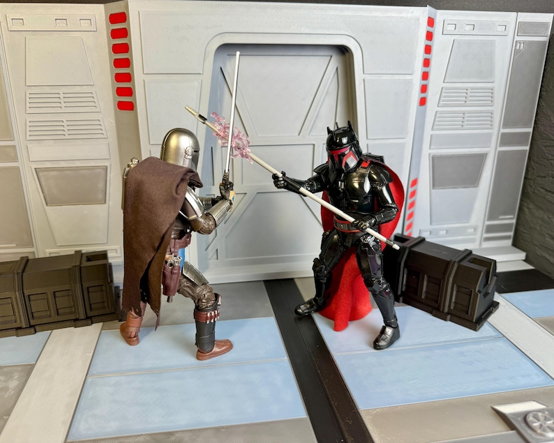 Puede incluir: Dos figuras de acci&oacute;n de Star Wars en una escena de batalla. Una figura es plateada y marr&oacute;n, con una capa y sosteniendo un bast&oacute;n blanco. La otra figura es negra y roja, tambi&eacute;n sosteniendo un bast&oacute;n blanco. El fondo es un interior gris y blanco.
