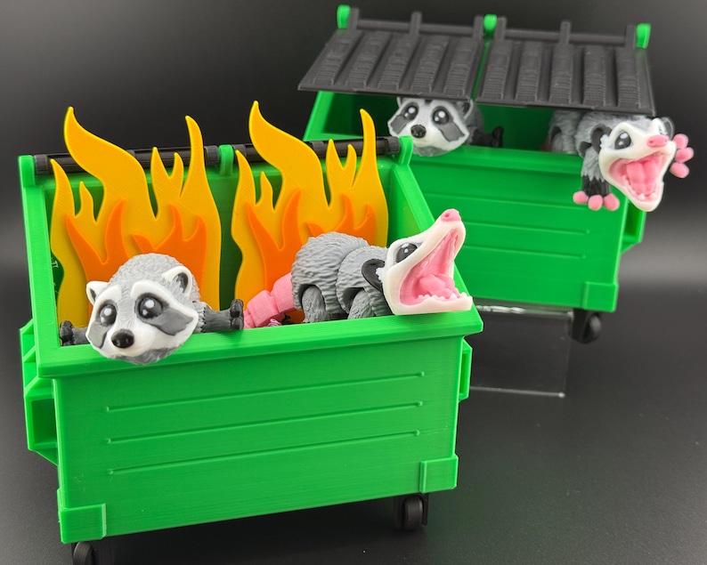 May include: Three raccoons are sitting in green wheelie bins. One wheelie bin has flames coming out of the top. The raccoons are all looking at the camera.