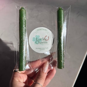 May include: Two slender, dark green ritual candles packaged in a clear plastic sleeve. A circular white label is centered between the candles, featuring a green clover, a horseshoe icon, and the text: Ritual Candle Luck Charm Increase your luck quickly.